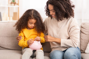 mother teaching young daughter early financial literacy by placing coins into piggy bank