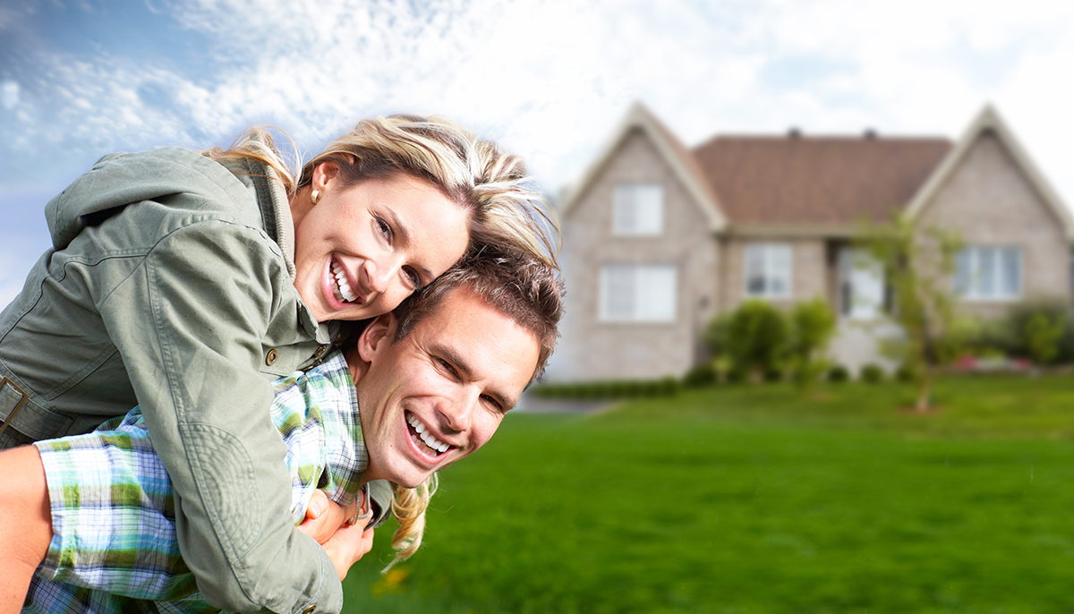 young man holding young woman on his back both smiling with their first new home in background